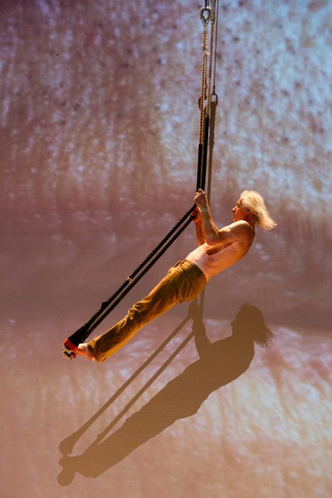 Mike Wright stands swinging on a trapeze, naked top half, white hair blowing behind him. 