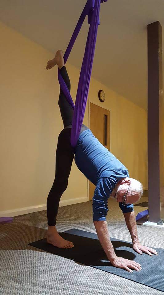 Guy is upside down, hands and one foot on the ground, the other suspended high in splits on aerial yoga hammock.
